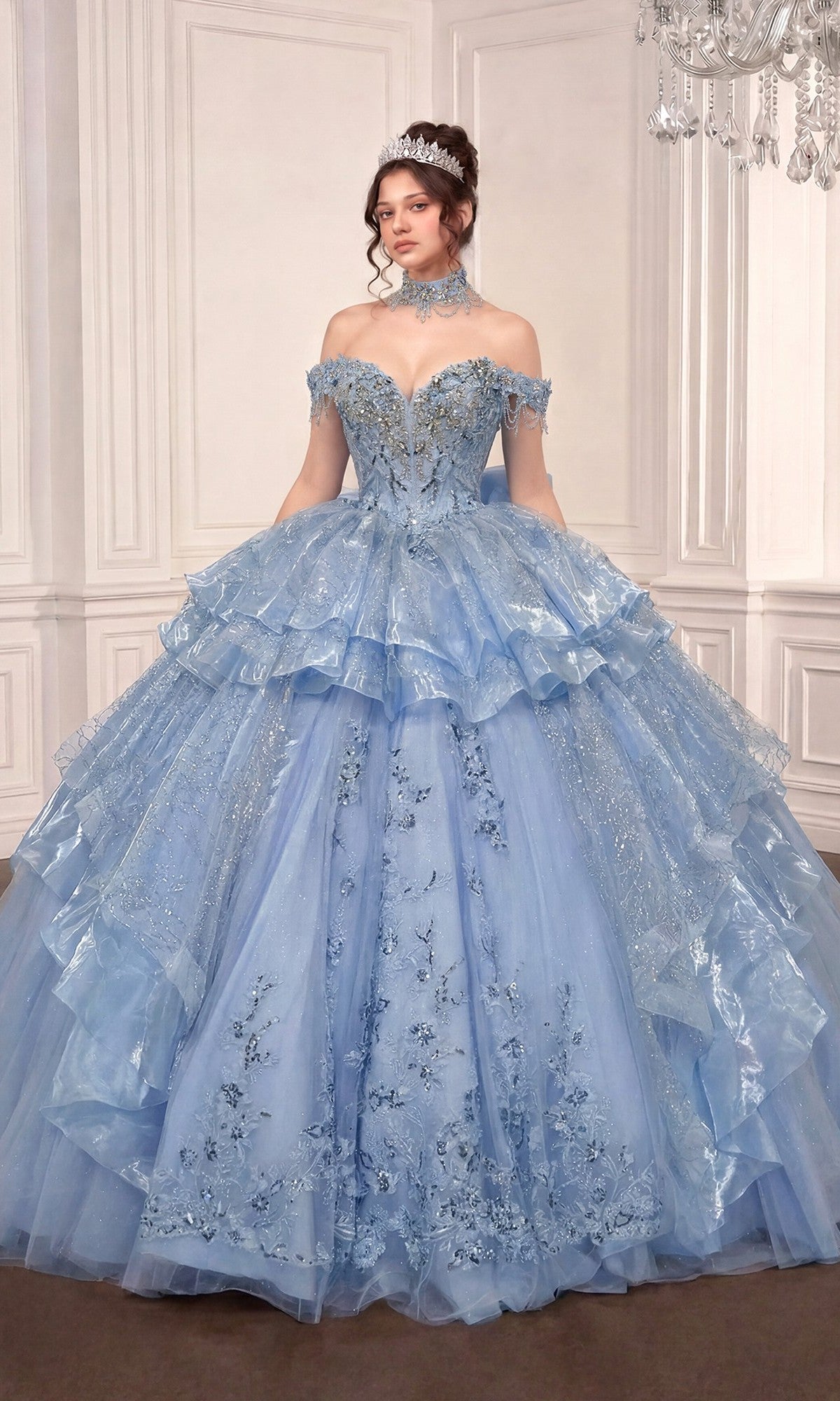 A woman in the Quinceanera Dress 15742 By Ladivine, a light blue gown with floral details and off-the-shoulder design, wears a tiara while standing in an elegant white-paneled room.